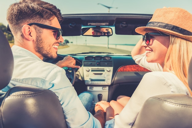 Enjoying their road trip. Side view of cheerful young couple holding hands and looking at each other while sitting inside of their convertible