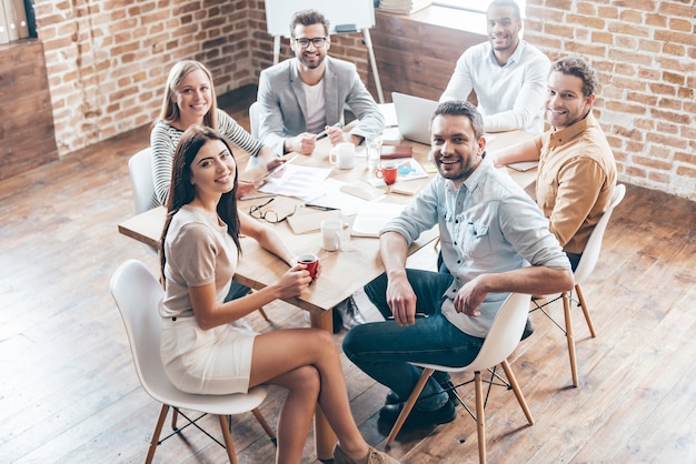 Enjoying their teamwork. Group of six cheerful young people looking at camera with smile while sitting at the table in office