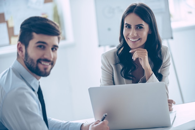 Enjoying their work together. Two cheerful coworkers looking at camera with smile while sitting at the office table