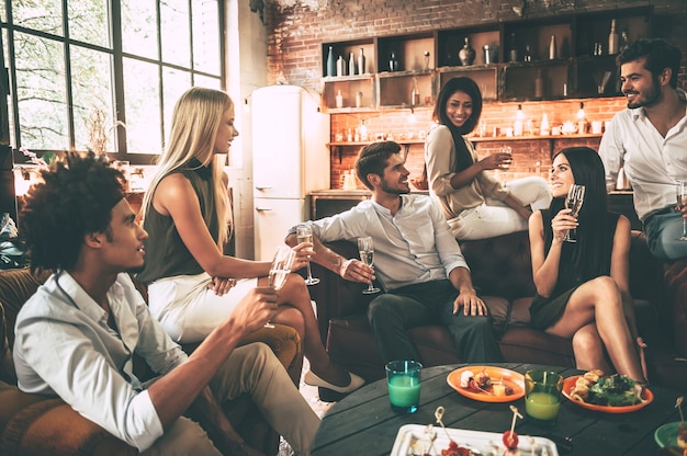 Enjoying time with friends. Group of cheerful young people enjoying food and drinks while spending nice time in cofortable chairs on the kitchen together