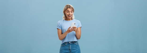 Entertained charming young european female with blond hair in casual outfit holding smartphone being