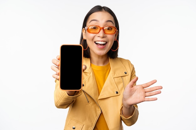 Enthusiastic asian female model showing smartphone app interface online store or website on mobile phone screen standing over white background