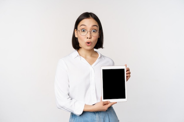 Enthusiastic asian woman office worker in glasses showing digital tablet screen demonstrating info on gadget display standing over white background