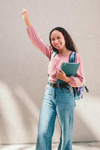 Enthusiastic black student woman raising her arm and holding her books against a wall. Education