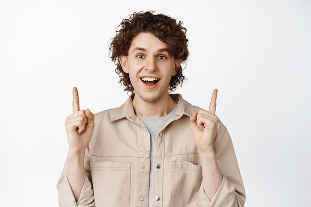 Enthusiastic curly young man pointing fingers up showing advertisement and smiling happy recommending store white background