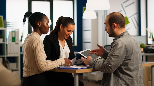 Enthusiastic diverse businesspeople reading annual financial report sitting at table in modern startup business office holding tablet and smiling. Team of multiethnic businesspeople working in company