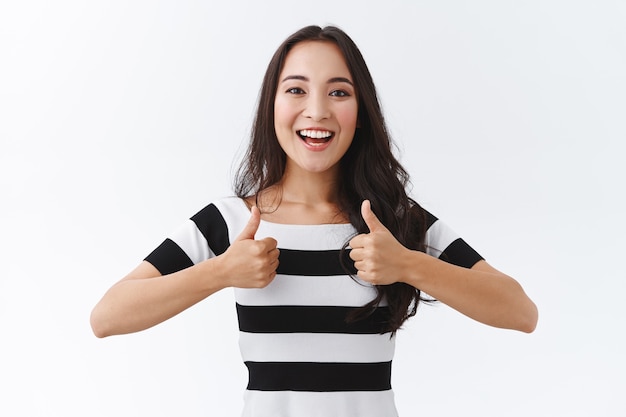 Enthusiastic girl encourage friend keep up. Attractive east-asian girl in striped t-shirt show thumbs-up and smiling excited, like awesome plan, approve great idea, stand white background