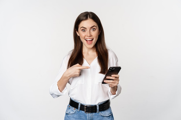 Enthusiastic girl pointing finger at smartphone and smiling showing big sale online using app on mob...