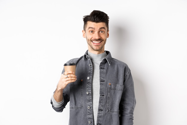 Enthusiastic handsome man holding coffee cup and smiling, drinking good drink from cafe takeout, standing against white background.