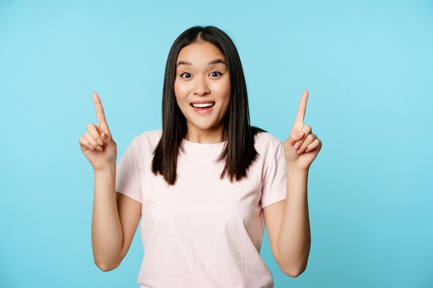 Enthusiastic korean girl shows advertisement points up and smiles amazed checks out promo deal stand...