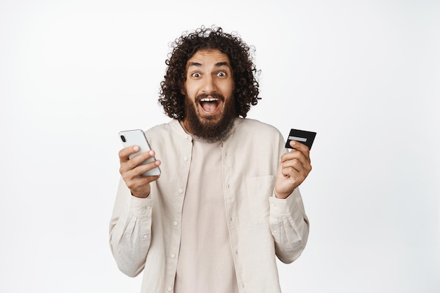 Enthusiastic male model screaming holding mobile phone and credit card reaction to online sale in shopping app white background