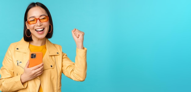 Enthusiastic smiling asian girl in sunglasses holding mobile phone and dancing laughing with smartphone standing over blue background