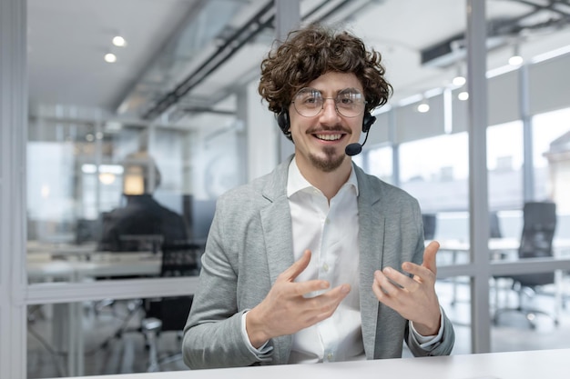 Enthusiastic young businessman with curly hair and headset communicates while working at his office