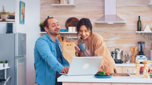 Entrepreneur working on laptop in kitchen and wife holding a cup of coffee looking at his work. Husband and wife cooking recipe food. Happy healthy together lifestyle. Family searching for online meal