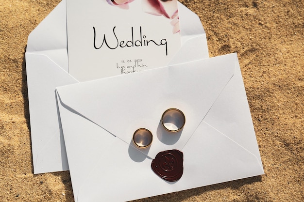 Envelopes with wedding invitations and gold rings on sandy beach top view