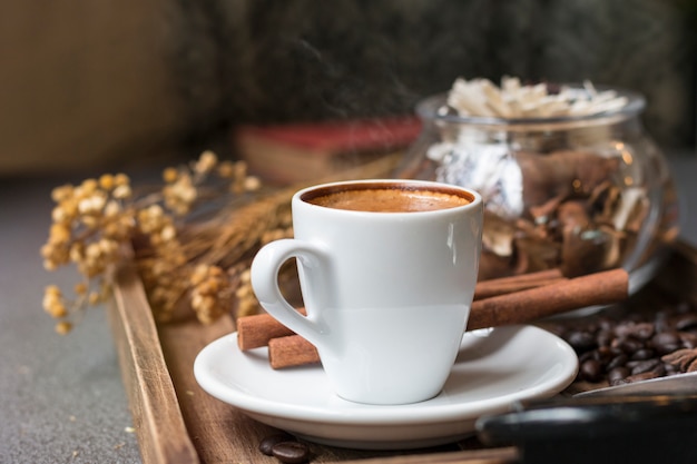 Espresso cup, coffee bean, cinnamon and dried flowers jar on wooden tray.