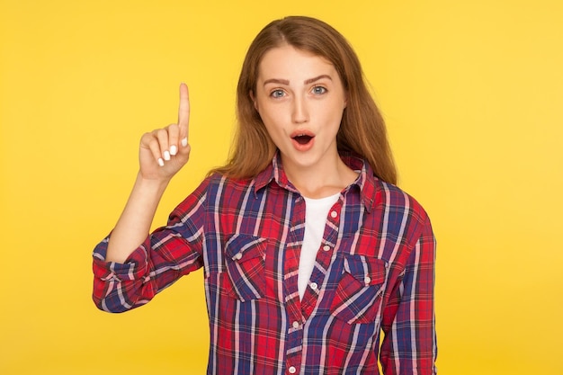 Eureka, excellent idea. Portrait of inspired smart ginger girl in checkered shirt pointing finger up and looking amazed at camera, got sudden solution, creative thoughts. studio shot yellow background