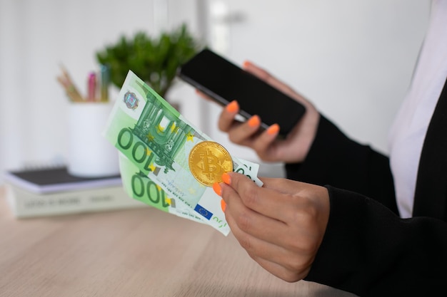 Euro bills and bitcoin at woman hands with phone in workplace