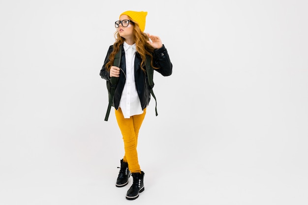 European attractive girl dressed in a yellow hat, glasses and a leather jacket with a backpack on his back in full growth onwhite wall with copyspace