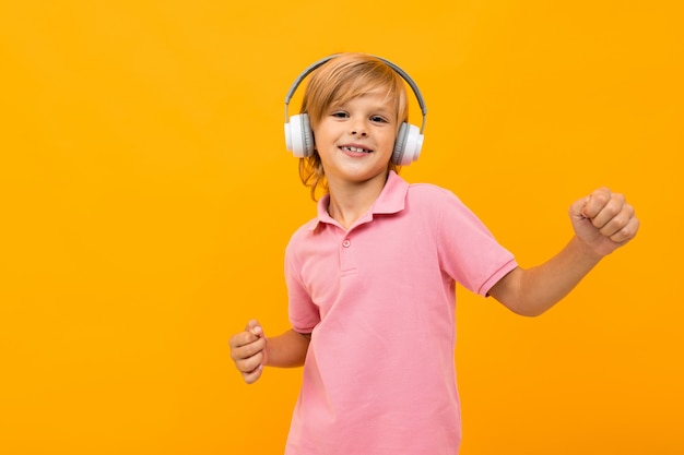 European blond boy in a pink T-shirt listens to music in white headphones on orange with copy space