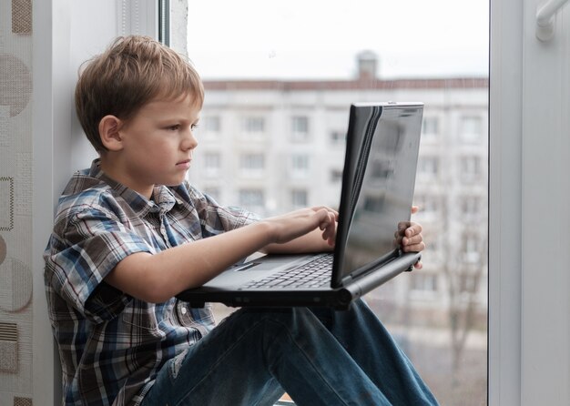 European boy sitting on the windowsill against the window with a black laptop