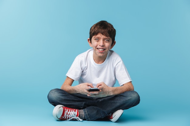 of european brunette boy  with freckles wearing white casual t-shirt holding and using smartphone while sitting on floor isolated over blue wall