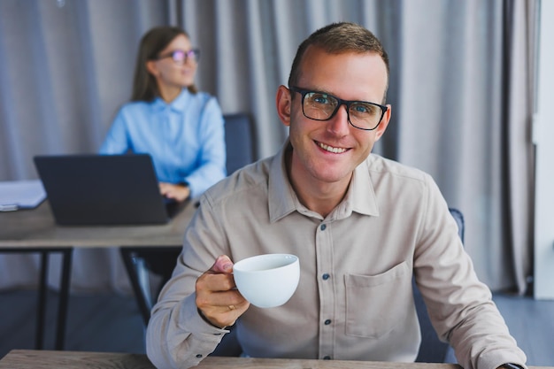 European businessman looking at something on laptop while working and drinking coffee The concept of a modern successful person Young guy in glasses sitting at the desk in the office with a laptop