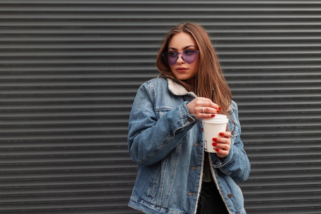 European cute young hipster woman in fashionable denim blue jacket in trendy purple glasses with cup of hot coffee is standing near a gray wall in the city. Pretty beautiful girl posing on the street.