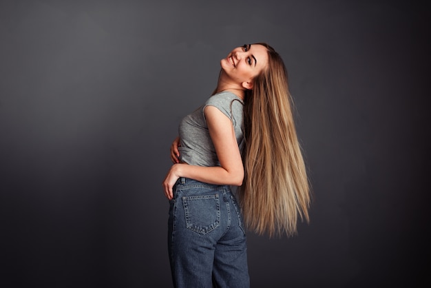 European girl in a gray top and jeans with blond long hair, stands sideways and holds her hand behind her head