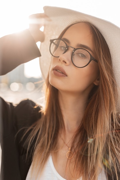 European lovely stylish young woman in vintage glasses in fashionable straw hat in black youth elegant blazer outdoors in city on sunny day. Vogue portrait beautiful girl in rays of summer bright sun.