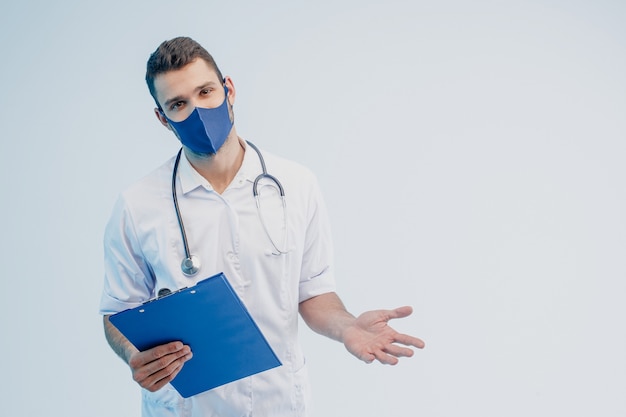 European male doctor in protective mask hold clipboard. Young man with stethoscope wearing white coat. Isolated on gray background with turquoise light. Studio shoot. Copy space.