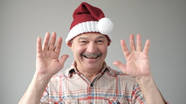 European pensioner in red santa claus hat waves hand in hello gesture