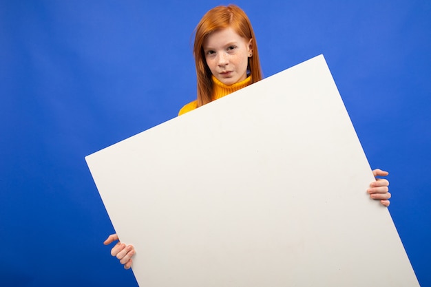 European red-haired teenage girl holding a white sheet of paper with a mockup for advertising on a blue studio background