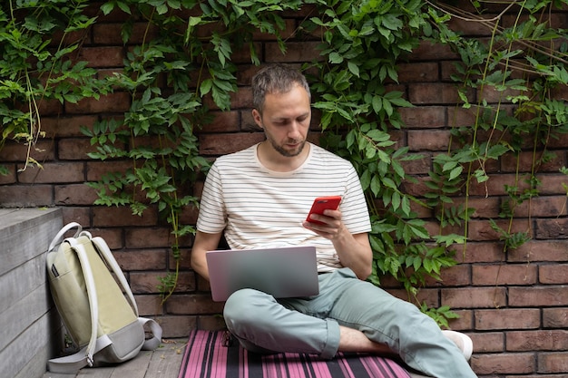 European student guy sitting with laptop outdoors using smartphone being distracted by mobile phone