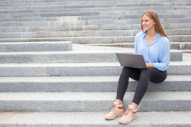 European student woman with happy face sitting on stairs and working on laptop looking away