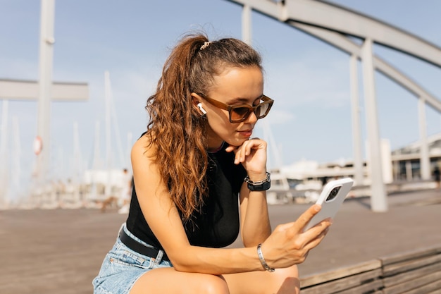 European stylish girl with wavy long hair wearing black top and shirts in sunglasses scrolling smartphone while sitting on the pier on background of yachts