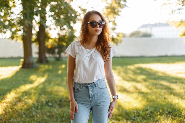 European stylish young hipster woman in trendy jeans in a white T-shirt in trendy sunglasses posing in a park among green trees