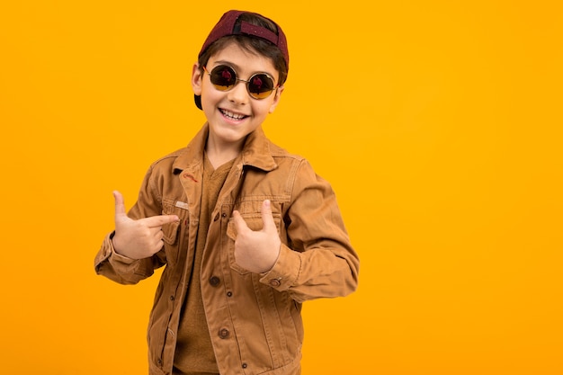 Photo european teenager boy in a casual stylish brown denim jacket and stylish glasses shows himself with his hand on a yellow background with copy space.