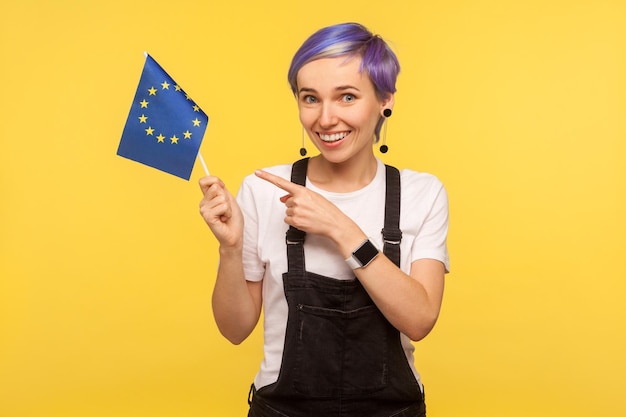 Photo european union flag portrait of positive hipster girl with violet short hair in denim overalls pointing at eu flag and looking at camera with toothy smile isolated on yellow background studio shot