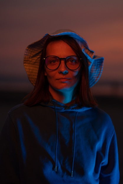 A european woman illuminated by neon light in a hat against the background of the sunset sky