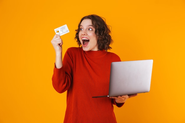 european woman wearing sweater using silver laptop and credit card while standing isolated on yellow