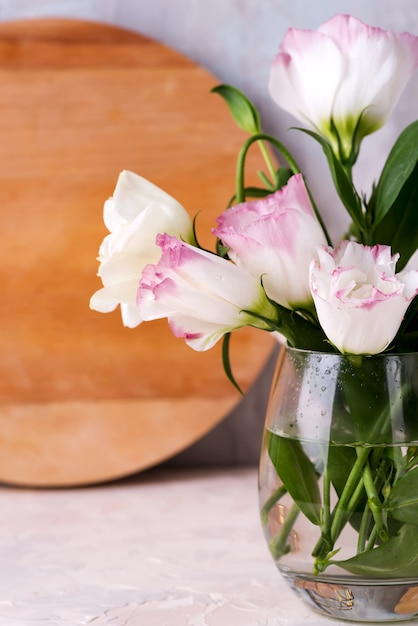 Eustoma flowers in vase on table near wooden plate and stone wall, close-up.