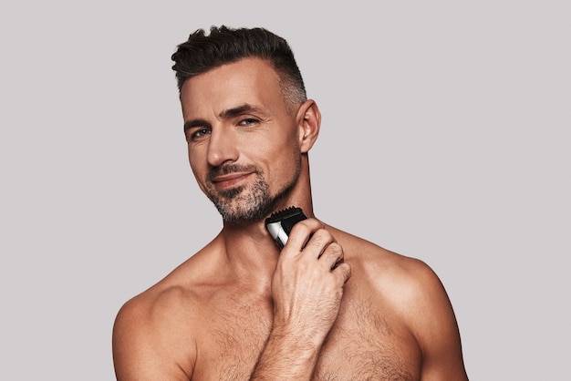 Everyday routine. Charming young man shaving and smiling while standing against grey background