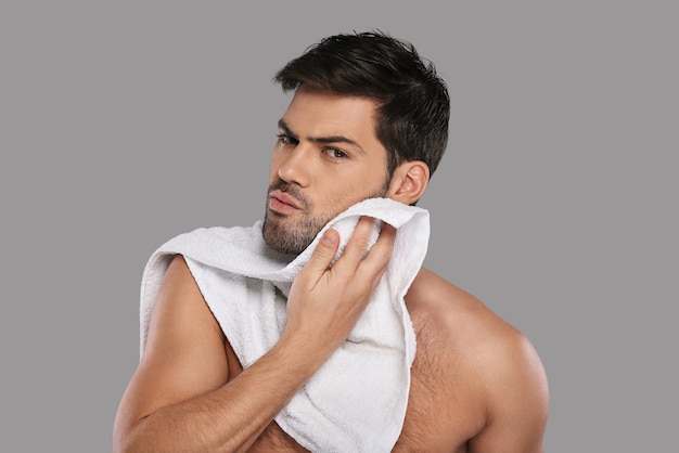 Everyday routine. Handsome young man wiping his face with towel while standing against grey background