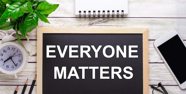 EVERYONE MATTERS written on a black background near pencils, a smartphone, a white notepad and a green plant in a pot