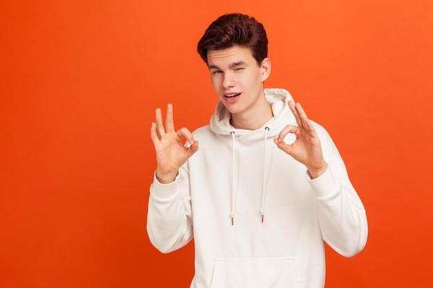 Everything is fine Stylish teenager in white hoody showing ok gesture and winking University application approval Indoor studio shot isolated on orange background