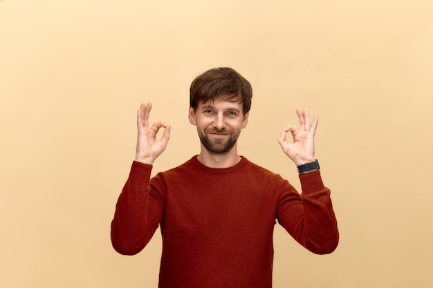 Everything is okay. Photo of young man with beard wearing sweater, show okay signs, has smile, posing against beige wall.