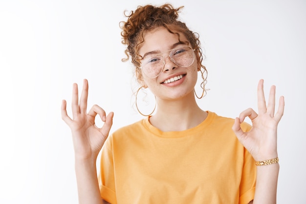 Everything super. Portrait charming chill confident happy smiling redhead girl wearing glasses orange t-shirt tilting head grinning self-assured show okay ok no problem gesture white background