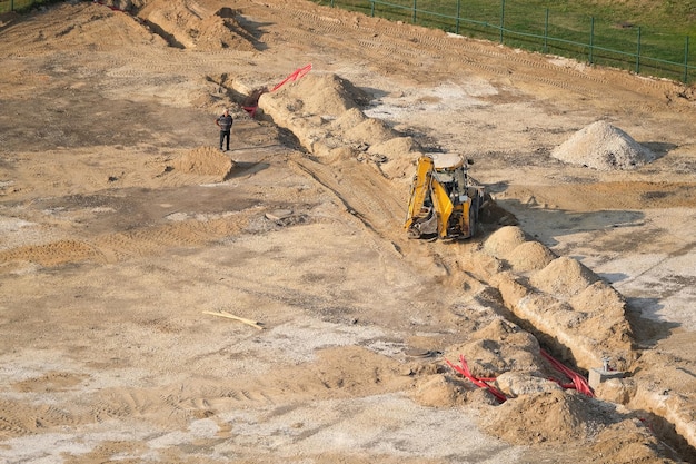 Excavator at the construction site dig trenches for cables and wires, while the work foreman stay ne