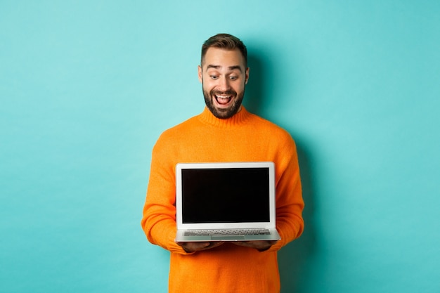 Excited adult male model showing laptop screen, demonstrating online promo, standing happy